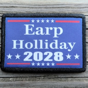 May include: A rectangular patch with a blue background and black border. The patch features the words "Earp Holliday 2028" in white, with red and white stars and stripes above and below the text. The patch is on a wooden surface.