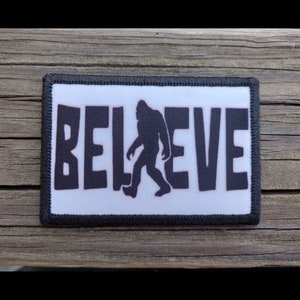 May include: A white patch with black stitching around the edges. The patch features a black silhouette of Bigfoot walking in front of the word "BELIEVE" in black letters.