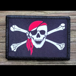 May include: A rectangular patch featuring a pirate flag design. The flag has a black background with a white skull wearing a red bandana and eye patch, and white crossed bones. The patch has a black border.