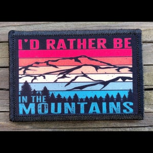 May include: Rectangular patch with the text "I'D RATHER BE IN THE MOUNTAINS" in white and blue. The patch features a mountain range design with a sunset color scheme of red, orange, and blue. The patch has a black border.
