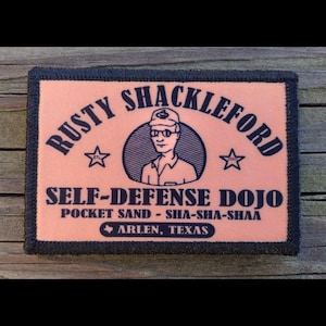 May include: A rectangular patch with the text "RUSTY SHACKLEFORD SELF-DEFENSE DOJO" and the phrase "POCKET SAND - SHA-SHA-SHAA". The patch features a cartoon illustration of a man in a hat and glasses, with two stars and the text "ARLEN, TEXAS".