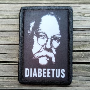 May include: A rectangular black patch with a white graphic illustration of a person wearing glasses and sporting a mustache. The text DIABEETUS is printed in white capital letters at the bottom of the patch, which features a black stitched border.