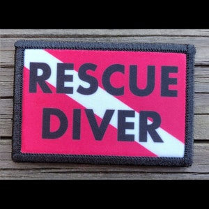 May include: Rectangular patch with a black border, featuring the words "RESCUE DIVER" in bold black letters. The background is bright pink with a white diagonal stripe. The patch is likely for a diving or rescue professional.