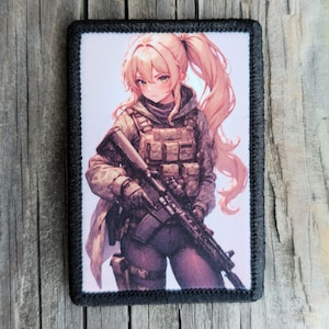May include: Rectangular patch featuring an anime-style illustration of a female figure in tactical gear, holding a rifle. The figure has long blonde hair and is set against a light background. The patch has a black border and is likely an accessory.