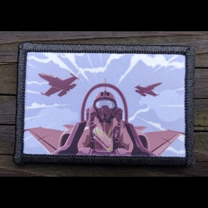 May include: A black and white embroidered patch with a pilot in a cockpit looking out at two fighter jets flying in formation against a blue sky with white clouds. The pilot is wearing a helmet and a brown flight suit.