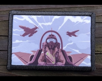 May include: A rectangular patch featuring a stylized illustration of a jet fighter pilot in a cockpit, with two jets flying in the sky. The patch has a black border and a light blue and purple color scheme.