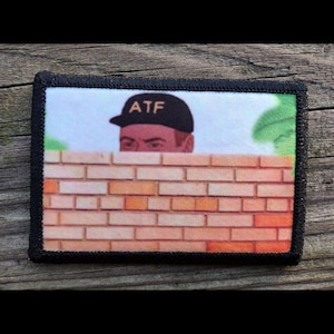 May include: A rectangular patch with a black border. The patch features a cartoon image of a person wearing a black cap with "ATF" printed on it, peeking over a brick wall. The wall is made of orange and brown bricks.