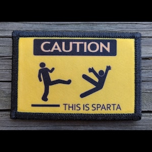 May include: A rectangular patch with a yellow background and black border. The patch features the word "CAUTION" above stick figures depicting a kick and a person falling. Below the figures, the text reads "THIS IS SPARTA."