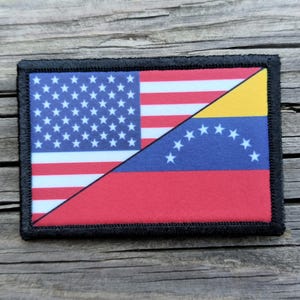 May include: A rectangular patch with a black border, featuring a split design. One half displays the American flag with stars and stripes, while the other half showcases the Venezuelan flag with stars and colors of yellow, blue, and red.