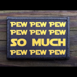May include: A rectangular patch with the words "PEW PEW PEW", "PEW PEW PEW", "SO MUCH", and "PEW PEW PEW" in yellow block letters on a black background. The patch has a black border and is likely made of fabric.