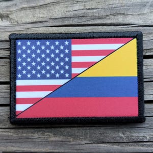 May include: Rectangular patch featuring a diagonal split design. The top left displays the United States flag with stars and stripes. The bottom right shows the flag of Colombia, with yellow, blue, and red stripes. The patch has a black border.