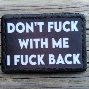 May include: Black rectangular patch with white text that reads "DON'T FUCK WITH ME I FUCK BACK". The patch has a black border and is displayed on a wooden surface.