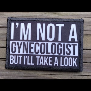 May include: Rectangular black and white patch with the text "I'M NOT A GYNECOLOGIST BUT I'LL TAKE A LOOK". The patch has a black border and is set against a wooden background.