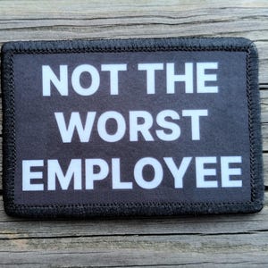 May include: A rectangular black patch with white text that reads "NOT THE WORST EMPLOYEE." The patch has a black border with stitched edges. The background is a weathered wooden surface.