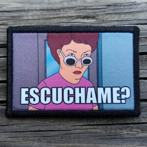 May include: Rectangular patch with a cartoon image of a woman wearing sunglasses and a pink shirt. The text "ESCUCHAME?" is printed in white. The patch has a black border.