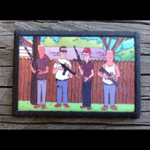 May include: A rectangular patch with a black border features a cartoon image of four men holding rifles. The men are standing in front of a wooden fence and a green lawn. The men are wearing casual clothing and holding drinks.