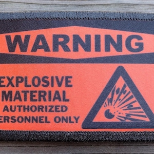 May include: A rectangular patch with a black border and a bright orange background. The patch features the word "WARNING" in black block letters, along with the words "EXPLOSIVE MATERIAL AUTHORIZED PERSONNEL ONLY" and a black triangle with an explosion symbol.