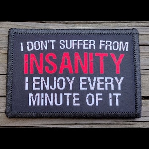 May include: A rectangular black patch with white text that reads "I DON'T SUFFER FROM INSANITY I ENJOY EVERY MINUTE OF IT." The word "INSANITY" is in red. The patch has a black border and is on a wooden surface.