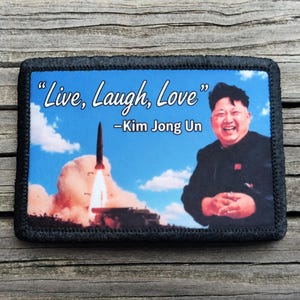 May include: Rectangular patch with a black border featuring a blue sky background. The patch includes the text "Live, Laugh, Love" and "-Kim Jong Un". A rocket launch and a portrait of Kim Jong Un are also visible.