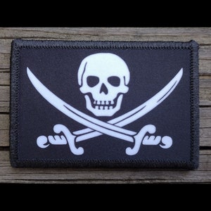 May include: A rectangular black patch featuring a white skull and crossbones design. Two white curved swords are crossed beneath the skull. The patch has a black stitched border, and is likely an emblem or accessory.