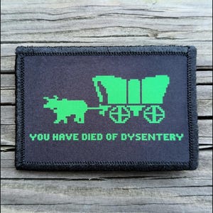 May include: A black rectangular patch with a green pixelated image of a covered wagon and oxen. The text "YOU HAVE DIED OF DYSENTERY" is below the image. The patch has a black border.