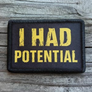 May include: Black rectangular patch with the words "I HAD POTENTIAL" in distressed yellow lettering. The patch has a black border and is set against a wooden background.