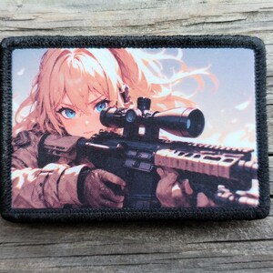 May include: A rectangular patch featuring an anime-style illustration of a person with blonde hair and blue eyes aiming a rifle with a scope. The patch has a black border and a detailed design.