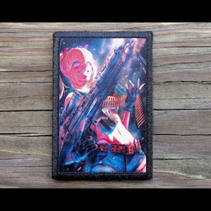 May include: A rectangular patch with a black border featuring a colorful anime-style illustration. The image depicts a smiling character holding a large firearm, with a red belt and blue pants. The background has a dark, smoky effect.