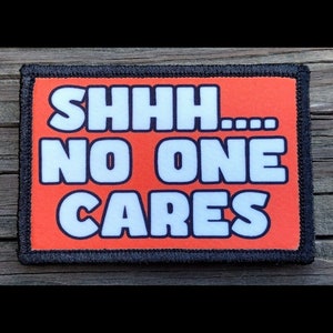 May include: Rectangular patch with the words "SHHH... NO ONE CARES" in white block letters on an orange background. The patch has a black border and is likely an iron-on or sew-on patch.