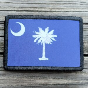 May include: A rectangular patch featuring the South Carolina state flag. The flag has a blue background with a white crescent moon in the upper left corner and a white palmetto tree in the center. The patch is bordered with black stitching.