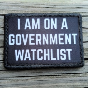 May include: A rectangular black patch with white text that reads "I AM ON A GOVERNMENT WATCHLIST." The patch has a black border with stitched edges. The background is a wooden surface.