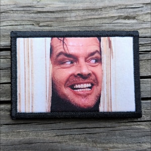 May include: Rectangular patch featuring a close-up of a man's face, peering through a door. The man has a wide, toothy grin and a crazed expression. The patch has a black border and is likely an iron-on or sew-on patch.
