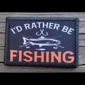 May include: A rectangular patch with the text "I'D RATHER BE FISHING" in white and orange. The patch features a white fish illustration with fishing rods and hooks on a black background, framed by a black border.