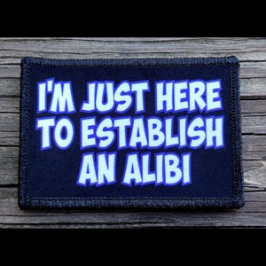 May include: A rectangular patch with the text "I'M JUST HERE TO ESTABLISH AN ALIBI" in white, outlined in blue, on a navy blue background. The patch has a black border and is set against a wooden surface.