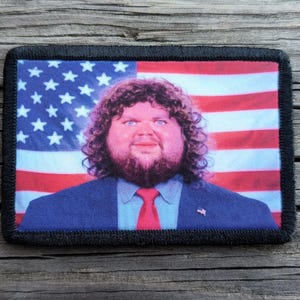 May include: Rectangular patch featuring a portrait of a man with a beard and curly hair, set against a backdrop of the American flag. The man is wearing a blue suit with a red tie. The patch has a black border.