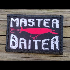 May include: Black rectangular patch with white text that reads "MASTER BAITER". A red fishing lure graphic is in the center. The patch has a black border and is likely for a jacket or bag.
