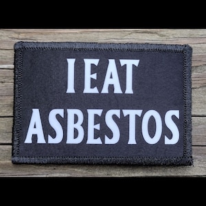 May include: Black rectangular patch with white text that reads "I EAT ASBESTOS". The patch has a black border with stitched edges. The background is a wooden surface.