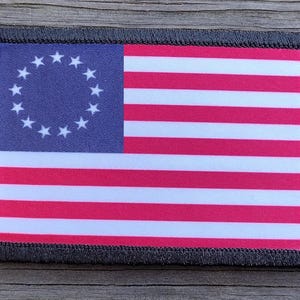 May include: A rectangular patch featuring the Betsy Ross flag design. The flag has a blue canton with 13 white stars arranged in a circle, and alternating red and white stripes. The patch is bordered with a black stitched edge.
