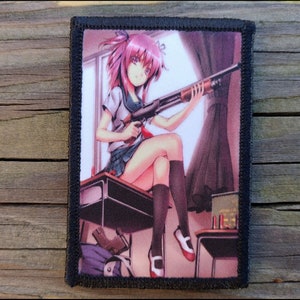 May include: A rectangular patch with a black border featuring an anime-style illustration. The image depicts a girl with pink hair holding a shotgun while sitting on a desk. The background includes a window, curtains, and a desk with ammunition.