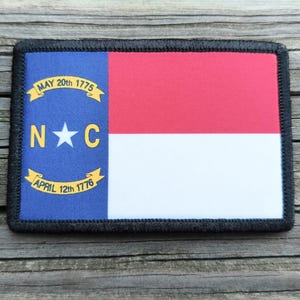 May include: A rectangular patch featuring the North Carolina state flag design. The flag has a blue vertical stripe with a white star and the letters "N" and "C", and the dates "May 20th 1775" and "April 12th 1776". The right side has a red stripe over a white stripe.