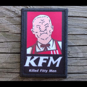 May include: A rectangular patch with a black border features a cartoon image of a stern-faced man in a white shirt, red apron, and bow tie, set against a red background. The text "KFM Killed Fifty Men" is displayed in white.