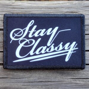 May include: A rectangular patch with the words "Stay Classy" in white cursive script on a navy blue background. The patch has a black border with a stitched edge, and is set against a wooden surface.