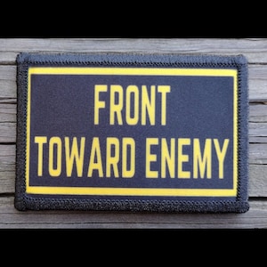May include: A rectangular patch with the text "FRONT TOWARD ENEMY" in yellow letters on a navy blue background. The patch has a yellow border and a black stitched edge.