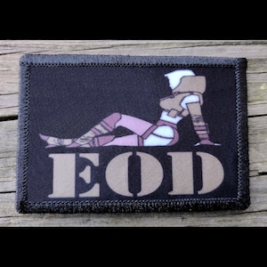 May include: Black rectangular patch with a stylized illustration of a figure in a futuristic outfit, in shades of brown, white, and pink. The text "EOD" is displayed in large, tan letters. The patch has a black border.