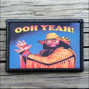 May include: Rectangular patch with a black border featuring a colorful image of a man in a cowboy hat and sunglasses. The text "OOH YEAH!" is in yellow. The man is wearing an orange sequined jacket.