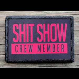 May include: A rectangular patch with the words "SHIT SHOW" in large, bright pink letters above "CREW MEMBER" in the same color. The text is on a dark gray background, framed by a black border.