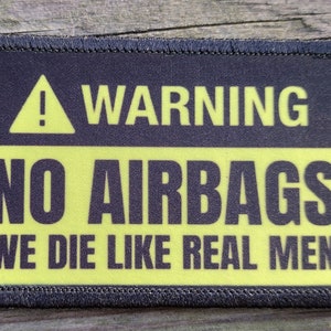 May include: A rectangular patch with a black border and a yellow and black design. The text reads "WARNING NO AIRBAGS WE DIE LIKE REAL MEN" with a yellow exclamation point symbol.