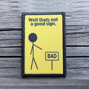 May include: A rectangular patch with a yellow background and a black border. The patch features a stick figure pointing towards a sign that reads "BAD". Above the figure, the text reads "Well thats not a good sign."
