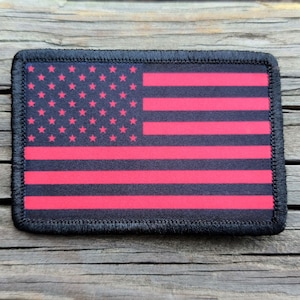 May include: A rectangular patch featuring the American flag design. The flag has a black background with red stripes and pink stars. The patch is bordered with black stitching.