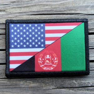 May include: Rectangular patch featuring the flags of the United States and Afghanistan. The US flag is on the left, with stars and stripes. The Afghan flag is on the right, with red, black, and green sections, and an emblem. The patch has a black border.